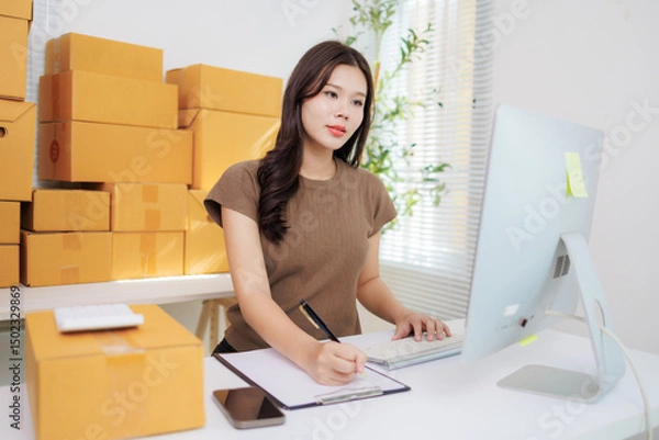 Fototapeta Young beautiful asian woman entrepreneur working from home checking orders and preparing packages for delivery, taking notes and using computer, surrounded by cardboard boxes