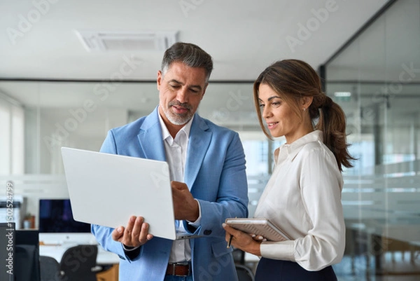 Fototapeta Busy professional smiling businessman and businesswoman leaders having conversation at work. Executives team of two business people working talking using laptop technology standing in corporate office