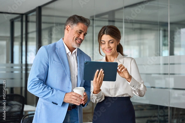 Obraz Two busy mature professional executives team business people talking using tablet computer having conversation standing in office, corporate managers working together with tab device.