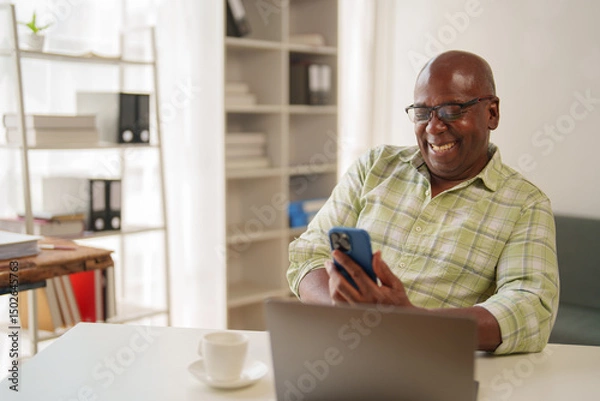 Fototapeta Happy african american man using mobile phone and smiling while working remotely from his home office, with laptop and cup of coffee on white desk