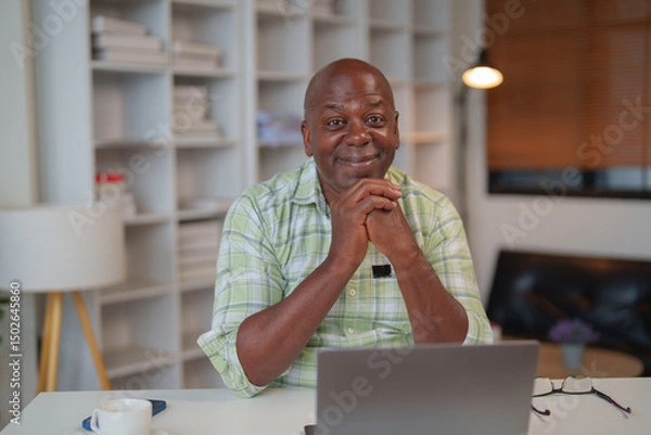 Fototapeta Happy african american businessman is sitting at desk and working remotely from home office using laptop, he is smiling and looking confident