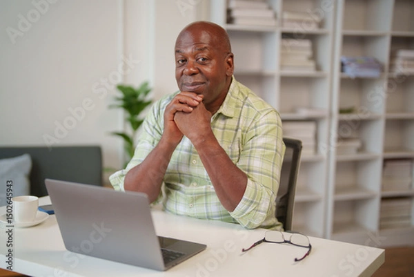 Fototapeta Smiling mature african american male freelancer sitting at desk with laptop computer, resting chin on clasped hands, working remotely from home, enjoying online freelance job
