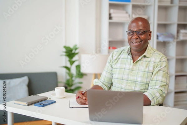 Fototapeta Senior black businessman working from his home office taking notes on a notepad and smiling while using his laptop, with a cup of coffee and a smartphone on his desk