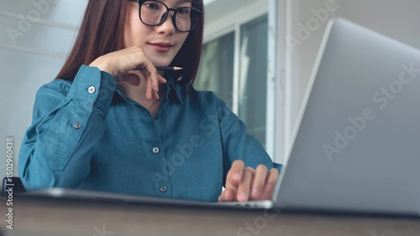 Obraz Young asian business woman working on laptop computer at office. Female student using laptop watching online webinar or training web course, looking at computer