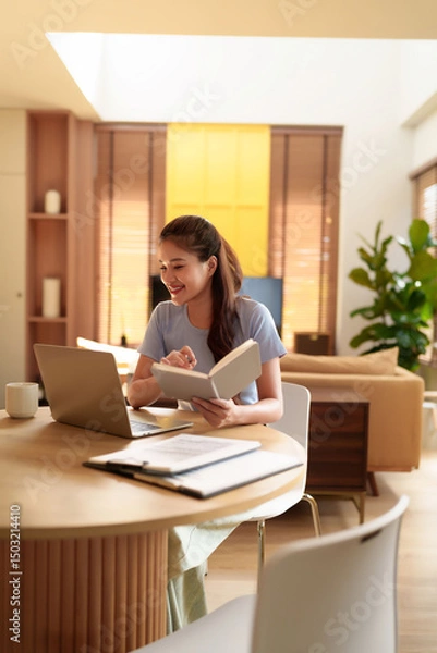 Obraz Young Asian woman working with laptop and book at home office. Happy female learning online, studying or distance education, remote worker in apartment.