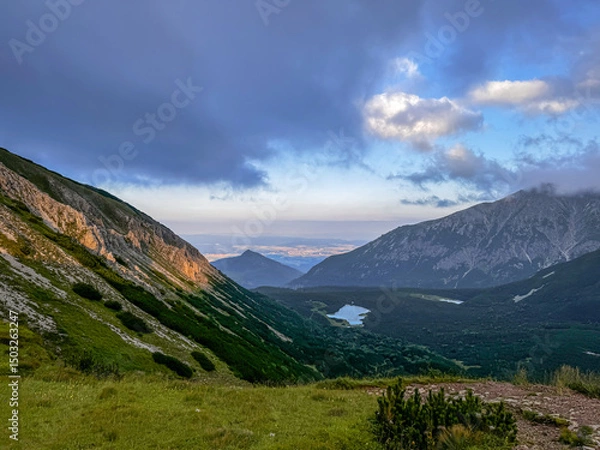 Fototapeta This breathtaking image captures a serene mountain valley landscape at twilight, showcasing lush hills, a reflective lake, and a dramatic cloud-filled sky