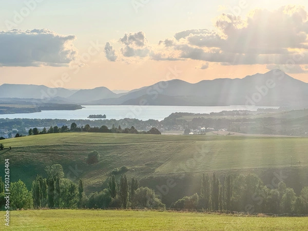 Fototapeta This stunning landscape features rolling hills, a serene lake, and majestic mountains, illuminated by the warm glow of the setting sun and soft rays of light breaking through clouds