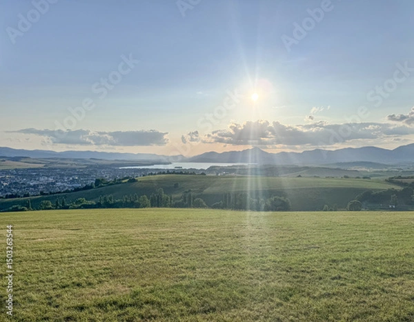 Fototapeta This stunning landscape features rolling hills, a serene lake, and majestic mountains, illuminated by the warm glow of the setting sun and soft rays of light breaking through clouds