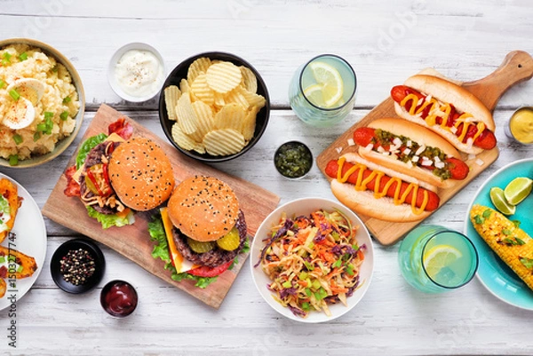 Obraz Summer food table scene with hot dogs, hamburger and sides. Above view over a white wood background.