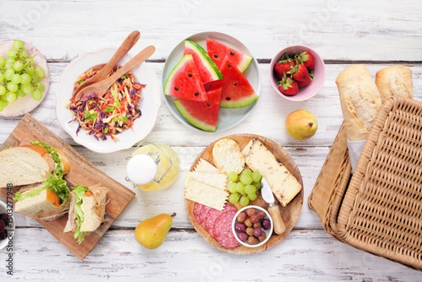Obraz Summer picnic food table scene. Top view over a white wood background. Picnic basket, fruit, salad, sandwiches and snacks.