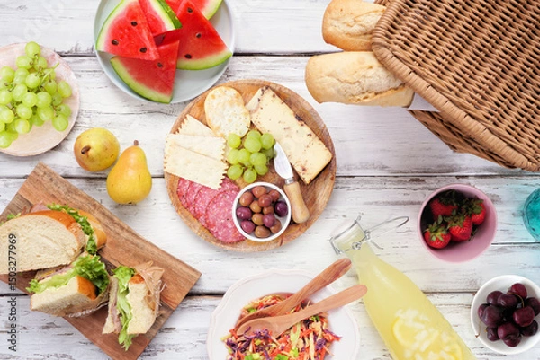 Obraz Summer picnic food table scene. Top view over a white wood background. Picnic basket, fruit, salad, sandwiches and snacks.
