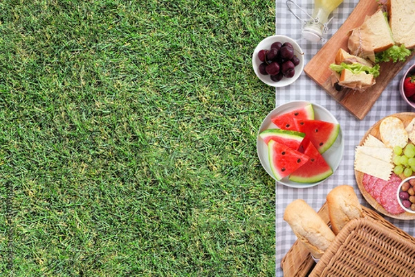 Obraz Summer picnic food side border. Top view with blanket over a green lawn background. Picnic basket, fruit, salad, sandwiches and snacks. Copy space.