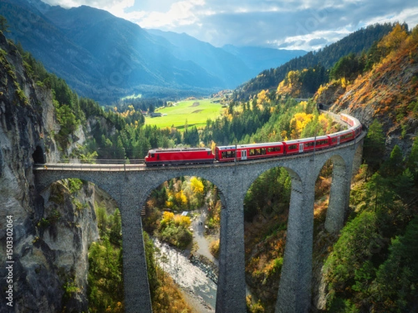 Obraz Aerial view of modern red train on Landwasser viaduct in alpine mountains, colorful forest at sunset in autumn. Bernina Express, Switzerland. Top view of train in Alps, railroad, green trees in fall