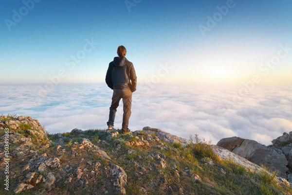 Obraz Man stands over the cover clouds on the edge of the abyss and looks on sun.