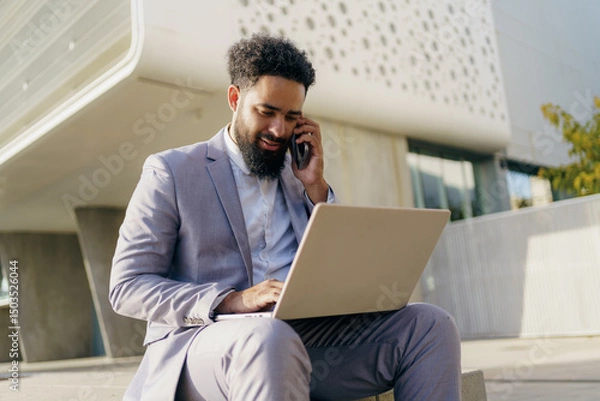 Obraz A professional man is utilizing a laptop while on the phone in a modern outdoor environment