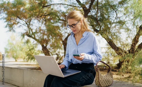 Obraz A businesswoman is working outdoors on her laptop while also utilizing her smartphone