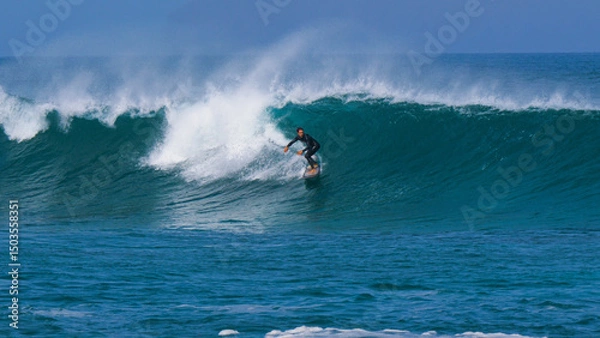 Fototapeta Regular footed rider surfing along a steep blue wave as offshore wind creates a dramatic spray. Powerful ocean swell forms perfect waves at surf spot on island of Lanzarote in the Canary Islands.