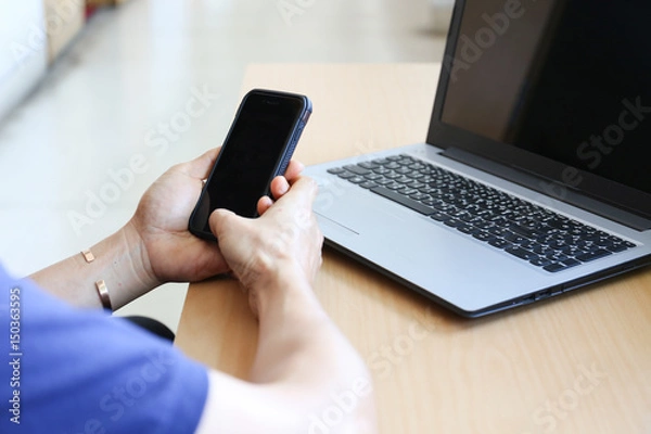 Fototapeta Man is holding mobile phone black display on hands in front of laptop on wooden desk