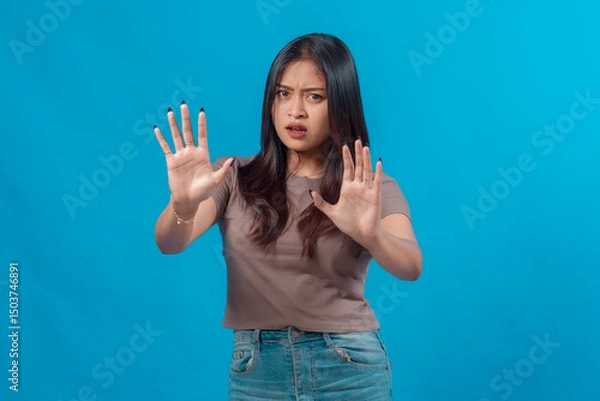 Fototapeta Young Asian woman looking concerned and raising both hands in a stop gesture, signaling rejection or warning, while standing in casual outfit against a plain blue background.