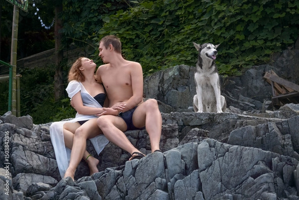 Fototapeta A young girl and a guy are sitting on the stone steps of a rock overgrown with vines, and they smile at each other, and a faithful dog is sitting next to them.