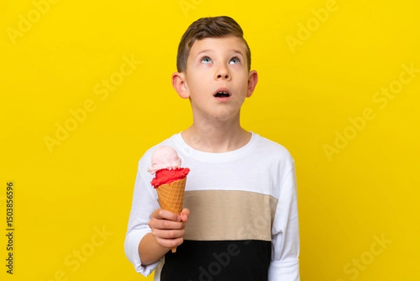 Fototapeta Little caucasian boy with a cornet ice cream isolated on yellow background looking up and with surprised expression
