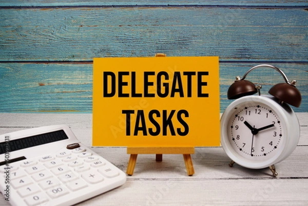 Fototapeta Delegate Tasks text written on paper card with calculator and alarm clock on wooden background