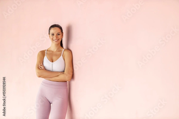 Fototapeta Portrait of fit proud sportswoman leaning on a wall with arms crossed and smiling at the camera.