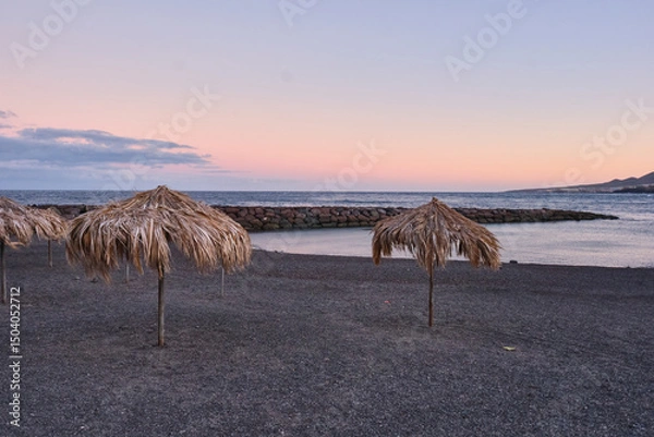 Fototapeta Tropical beach with straw umbrellas at sunset, volcanic sand shore
