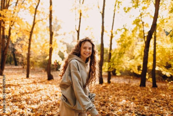 Fototapeta Smiling young woman enjoys the autumn weather in the forest with the yellow leaves at sunset