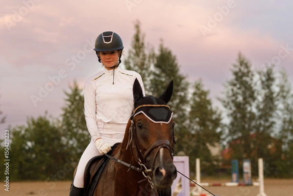 Fototapeta Front view of chestnut horse in harness with female rider jockey in hat helmet and white uniform with bridle stack and boots