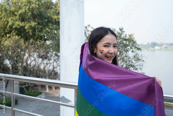 Fototapeta A joyful young woman proudly displays a rainbow flag, symbolizing love and diversity, with a river backdrop. Celebrating inclusivity and unity.