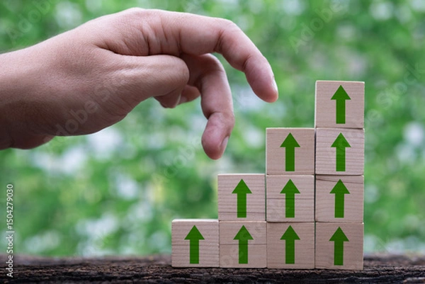 Fototapeta A hand reaches towards stacked blocks with green arrows, symbolizing growth and progress against a natural backdrop.