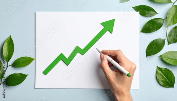 Fototapeta Green arrow graph eco investing stocks concept with hand holding pen and leaves on blue background for growth and success