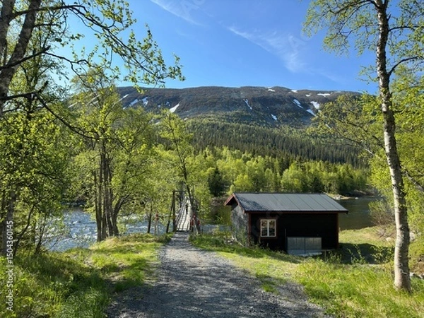 Obraz Cabin nestled near a river with a suspension bridge and a mountain backdrop in north Sweden.