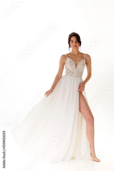 Fototapeta Elegant model poses in flowing white dress with intricate details and high slit against bright background