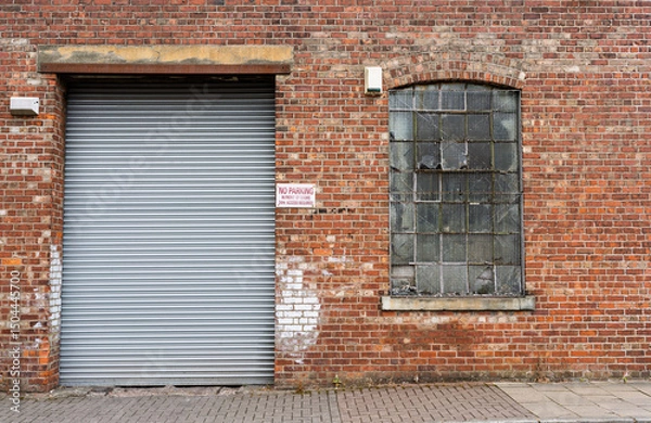 Fototapeta Brick warehouse facade with large metal shutter door and worn window on an industrial building.