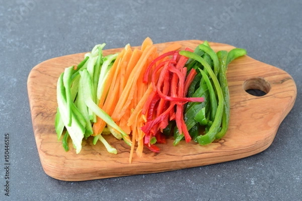 Fototapeta Fresh cucumber, carrot, red and green sweet paprika sliced in stripes on an olive wood cutting board on a grey abstract background. Step by step cooking
