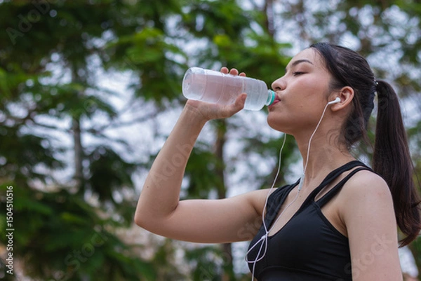 Obraz Portrait of beautiful Asian runner drinking water from plastic bottle because she feels tired from run and wants take break drink water. Beautiful Asian woman taking break drink water after tiring run
