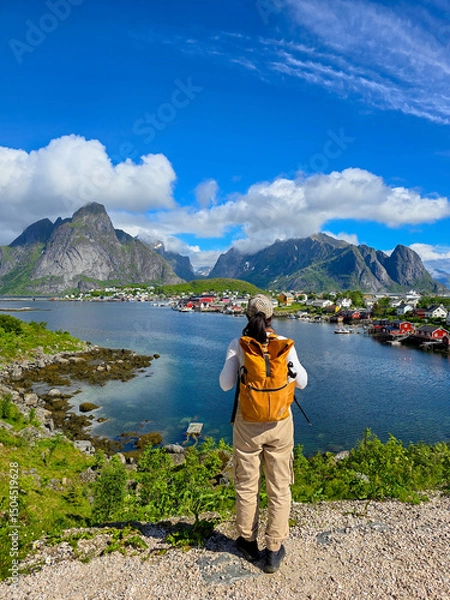 Fototapeta A traveler stands by the serene waters of a Norwegian fjord, surrounded by stunning mountains and charming fishing villages. The bright sky adds vibrancy to this enchanting location. Lofoten Norway