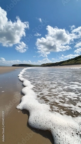 Fototapeta Gentle ocean waves roll steadily over a sandy beach beneath a radiant, sunny sky. Vertical.