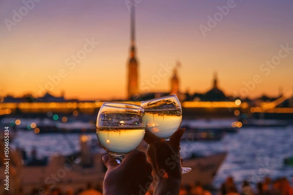 Fototapeta close-up view of two hands clinking glasses of champagne, Neva River and Peter and Paul Fortress are reflected upside down in glass, which are blurred in background, at evening, St Petersburg, Russia