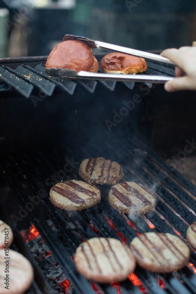 Fototapeta Juicy burger patties sizzling on a charcoal grill with glowing embers below. A hand uses tongs to toast burger buns on the upper rack. Backyard barbecue cooking in progress.