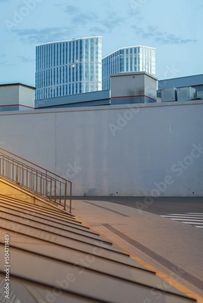 Fototapeta Minimalist cityscape with tall modern glass towers behind a plain concrete wall at sunset. Urban contrast between architecture and infrastructure in soft evening light.