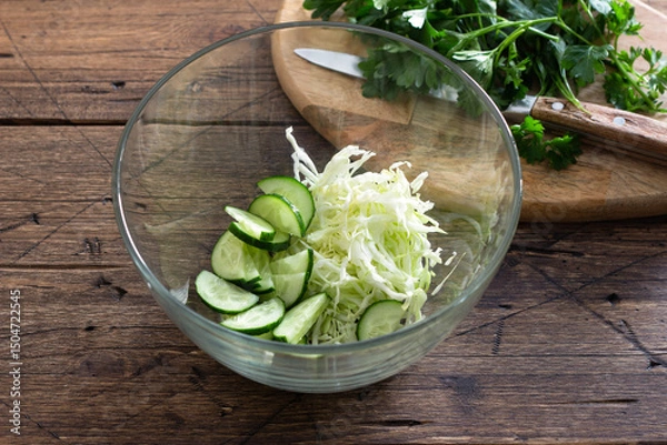 Obraz Fresh cucumber and cabbage salad in glass bowl on wooden table, healthy vegetarian food preparation concept
