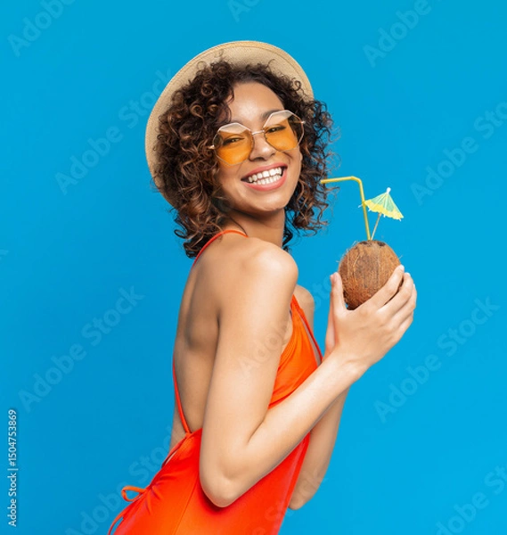 Obraz Happy black girl in swimsuit enjoying exotic summer coconut cocktail and smiling at camera, posing over blue background in studio, copy space