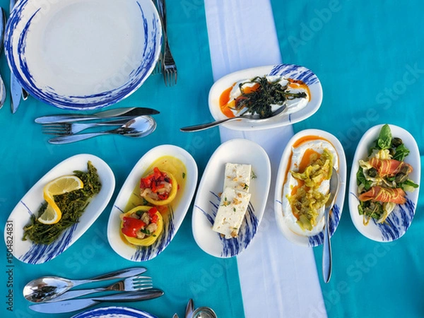 Fototapeta Assortment of Mediterranean appetizers served on blue tablecloth. Various dishes with vegetables and cheeses in oval plates, ready for eat. Top view. 