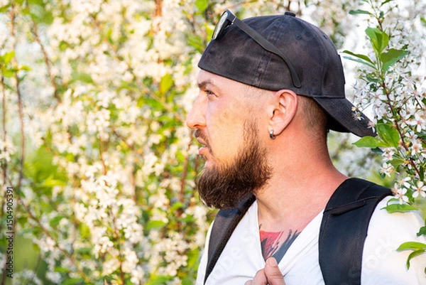 Fototapeta Portrait of Man with Beard in Baseball Cap Against White Flowery Tree in Spring.