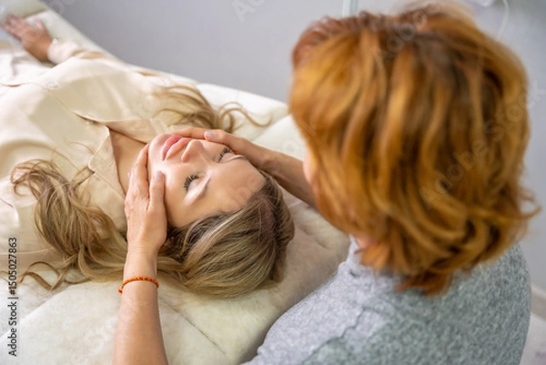 Obraz Female practitioner performs facial lifting technique on a young woman lying on a couch. Concept of energy healing, face therapy and alternative wellness practice from Access Bars.