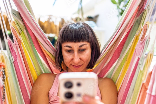 Fototapeta Woman lying in hammock using smartphone. summer mood. girl relaxing swinging in hammock and scrolling social media near summer camping.