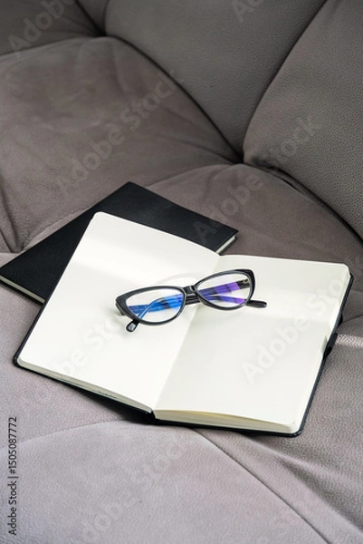 Fototapeta Pair of Stylish Black Glasses Rests on an Open Notebook Placed on Soft Gray Couch.